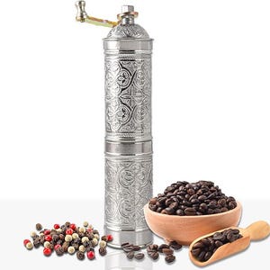 Pode incluir: Um moedor de caf&eacute; manual prateado com um design floral ornamentado, uma tigela de madeira cheia de gr&atilde;os de caf&eacute; e uma colher de madeira. H&aacute; tamb&eacute;m uma pequena pilha de gr&atilde;os de pimenta.