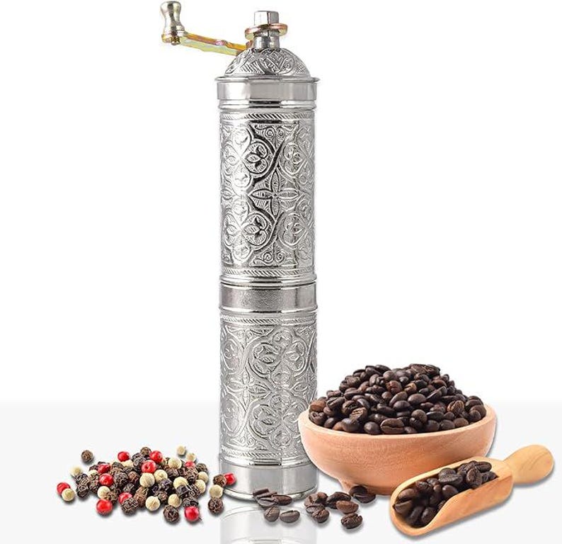 Pode incluir: Um moedor de caf&eacute; manual prateado com um design floral ornamentado, uma tigela de madeira cheia de gr&atilde;os de caf&eacute; e uma colher de madeira. H&aacute; tamb&eacute;m uma pequena pilha de gr&atilde;os de pimenta.