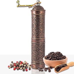 Pode incluir: Um moedor de caf&eacute; manual de cor cobre com um design floral ornamentado. O moedor tem uma manivela e est&aacute; ao lado de uma tigela de madeira com gr&atilde;os de caf&eacute;, uma colher de madeira com gr&atilde;os de caf&eacute; e uma pilha de gr&atilde;os de pimenta.