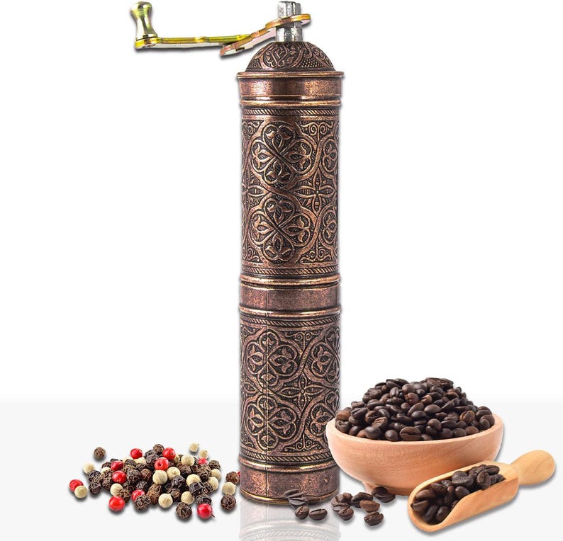 Pode incluir: Um moedor de caf&eacute; manual de cor cobre com um design floral ornamentado. O moedor tem uma manivela e est&aacute; ao lado de uma tigela de madeira com gr&atilde;os de caf&eacute;, uma colher de madeira com gr&atilde;os de caf&eacute; e uma pilha de gr&atilde;os de pimenta.