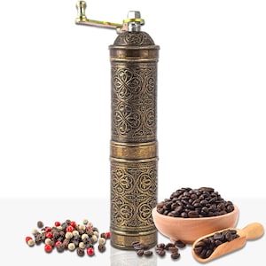 Puede incluir: Un molinillo de café manual de estilo antiguo con un diseño ornamental en relieve en tonos bronce y oro. Junto a él, granos de café enteros en un cuenco de madera pequeño y una cuchara de madera, junto con un montón de granos de pimienta.