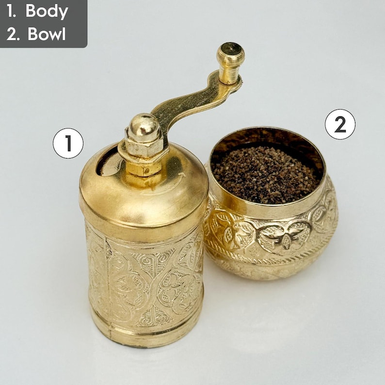 May include: A gold-colored pepper grinder and bowl set. The grinder has a cylindrical body with an ornate, engraved pattern. The bowl is filled with dark peppercorns and has a matching engraved design. The grinder features a crank handle.
