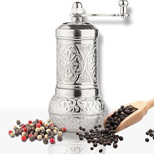 May include: A silver-colored pepper mill with an ornate floral design. The mill has a crank handle and sits next to a wooden scoop filled with black peppercorns. A small pile of mixed peppercorns is also visible.