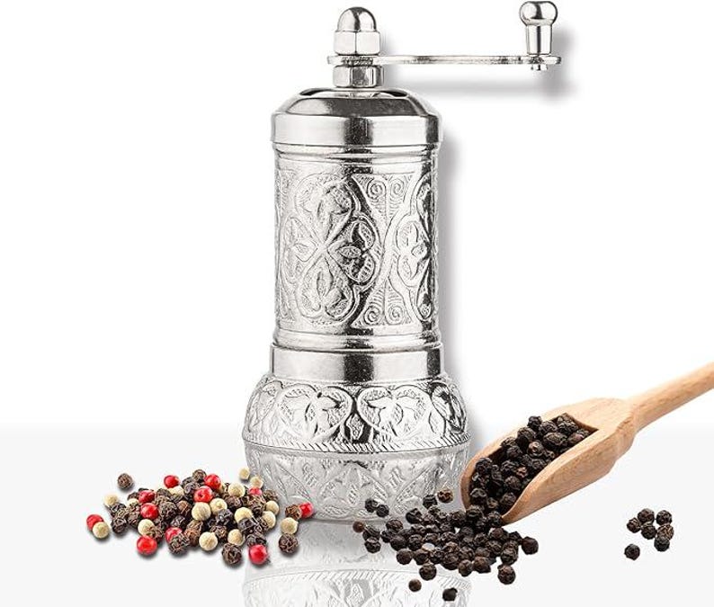 May include: A silver-colored pepper mill with an ornate floral design. The mill has a crank handle and sits next to a wooden scoop filled with black peppercorns. A small pile of mixed peppercorns is also visible.