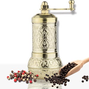 May include: A gold-colored pepper mill with an ornate, engraved design. The mill has a crank handle and is accompanied by a wooden scoop filled with black peppercorns and a pile of mixed peppercorns.