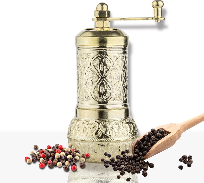 May include: A gold-colored pepper mill with an ornate, engraved design. The mill has a crank handle and is accompanied by a wooden scoop filled with black peppercorns and a pile of mixed peppercorns.