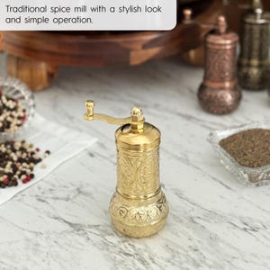 May include: A gold-colored spice mill with an ornate, engraved design. The mill has a hand crank and is sitting on a marble surface. The text in the image reads: "Traditional spice mill with a stylish look and simple operation."