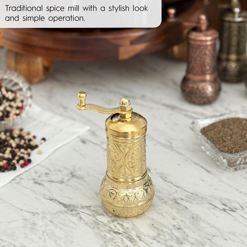 May include: A gold-colored spice mill with an ornate, engraved design. The mill has a hand crank and is sitting on a marble surface. The text in the image reads: "Traditional spice mill with a stylish look and simple operation."