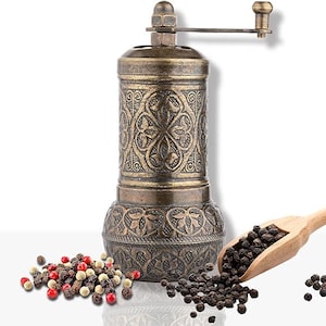 May include: An antique-style pepper mill with an ornate, embossed design in bronze. A wooden scoop holds black peppercorns, with a scattering of peppercorns in various colors nearby. The mill has a hand crank and a classic aesthetic.