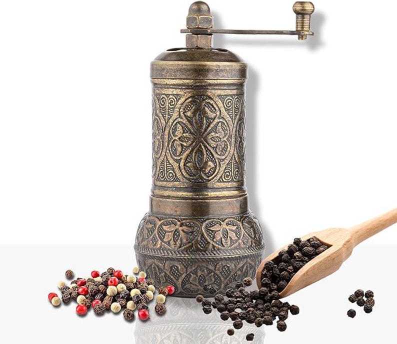 May include: An antique-style pepper mill with an ornate, embossed design in bronze. A wooden scoop holds black peppercorns, with a scattering of peppercorns in various colors nearby. The mill has a hand crank and a classic aesthetic.