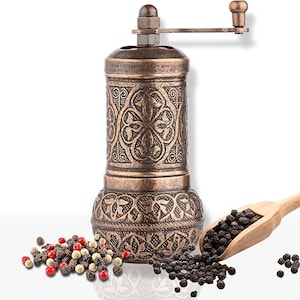 May include: A bronze-colored pepper mill with an ornate floral design. A wooden scoop holds black peppercorns, and a small pile of mixed peppercorns sits nearby. The mill has a hand crank and is designed for grinding spices.