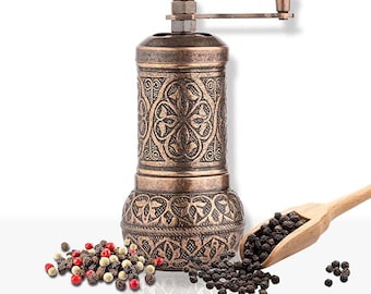 Pode incluir: Um moedor de pimenta bronzeado com um design floral ornamentado. Uma colher de madeira contém grãos de pimenta preta, e uma pequena pilha de pimenta mista está por perto. O moedor tem uma manivela para moer especiarias.