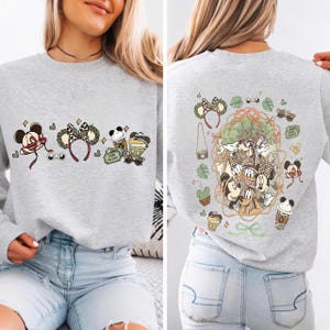 May include: A light gray sweatshirt featuring Mickey Mouse and Minnie Mouse designs. The front has several cartoon images, including a Mickey Mouse head with a bow, a headband with ears, and a coffee cup. The back has a larger image of the characters.