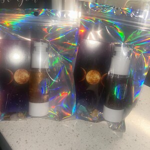 K&ouml;nnte beinhalten: Zwei Kosmetikflaschen mit wei&szlig;en Pumpen und goldglitzerndem Inhalt sind in schillernden Zellophanbeuteln verpackt. Die Beutel haben ein Hintergrundbild eines Himmelsdesigns mit Mondphasen. Der Text "st God" ist im Hintergrund sichtbar.