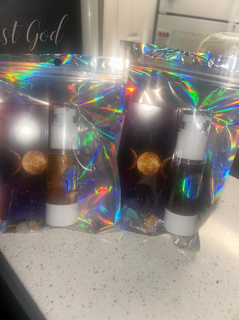 K&ouml;nnte beinhalten: Zwei Kosmetikflaschen mit wei&szlig;en Pumpen und goldglitzerndem Inhalt sind in schillernden Zellophanbeuteln verpackt. Die Beutel haben ein Hintergrundbild eines Himmelsdesigns mit Mondphasen. Der Text "st God" ist im Hintergrund sichtbar.