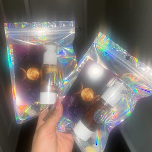 K&ouml;nnte beinhalten: Zwei transparente Flaschen mit wei&szlig;en Pumpen und goldener Fl&uuml;ssigkeit im Inneren, verpackt in schillernden Beuteln. Jeder Beutel zeigt ein Himmelsdesign mit Mond und Sternen. Die Flaschen werden in einer Hand gehalten.