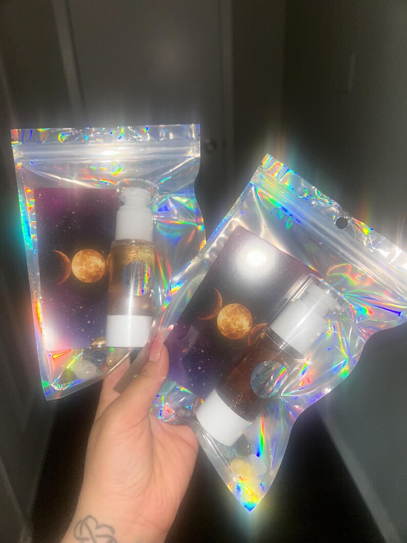 K&ouml;nnte beinhalten: Zwei transparente Flaschen mit wei&szlig;en Pumpen und goldener Fl&uuml;ssigkeit im Inneren, verpackt in schillernden Beuteln. Jeder Beutel zeigt ein Himmelsdesign mit Mond und Sternen. Die Flaschen werden in einer Hand gehalten.