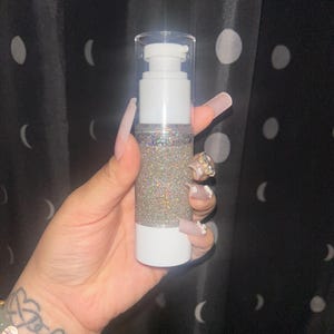 K&ouml;nnte beinhalten: Eine wei&szlig;e und transparente Pumpflasche gef&uuml;llt mit schimmerndem Glitzer. Die Flasche hat einen wei&szlig;en Boden und einen transparenten Deckel mit einem Pumpspender. Der Glitzer im Inneren reflektiert mehrere Farben. Der Hintergrund ist ein schwarzer Vorhang mit wei&szlig;en Punkten.