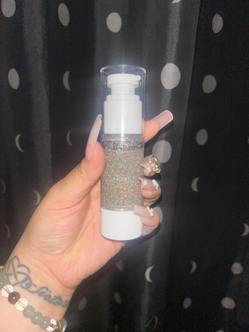 K&ouml;nnte beinhalten: Eine wei&szlig;e und transparente Pumpflasche gef&uuml;llt mit schimmerndem Glitzer. Die Flasche hat einen wei&szlig;en Boden und einen transparenten Deckel mit einem Pumpspender. Der Glitzer im Inneren reflektiert mehrere Farben. Der Hintergrund ist ein schwarzer Vorhang mit wei&szlig;en Punkten.