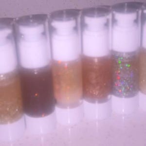 K&ouml;nnte beinhalten: Acht transparente Flaschen mit wei&szlig;en Pumpen, jede gef&uuml;llt mit einer anderen Farbe Glitzer. Die Farben reichen von Orange bis Braun, mit goldenen und irisierenden Glitzeroptionen. Die Flaschen sind in einer Reihe angeordnet.