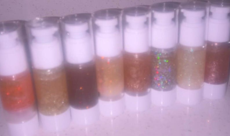 K&ouml;nnte beinhalten: Acht transparente Flaschen mit wei&szlig;en Pumpen, jede gef&uuml;llt mit einer anderen Farbe Glitzer. Die Farben reichen von Orange bis Braun, mit goldenen und irisierenden Glitzeroptionen. Die Flaschen sind in einer Reihe angeordnet.