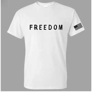 May include: White t-shirt with the word "FREEDOM" printed in black across the chest. An American flag graphic is on the left sleeve. The shirt is a classic crew neck style.