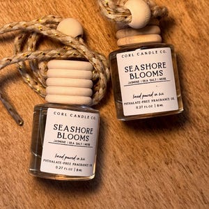 May include: Two small glass bottles with wooden tops and hanging cords. The labels read "SEASHORE BLOOMS" with scent notes and "hand poured in wi". Each bottle contains 8 ml of phthalate-free fragrance oil.