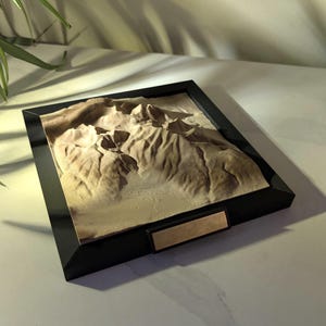 Puede incluir: Un mapa de relieve topográfico 3D cuadrado con marco negro que presenta un paisaje montañoso texturizado de color tostado. El marco incluye una pequeña placa metálica rectangular en blanco centrada en el borde inferior.