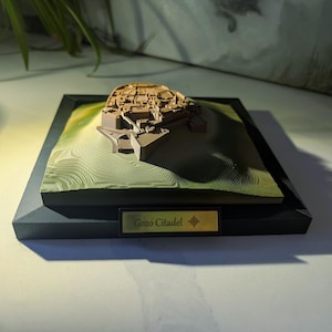 May include: A 3D topographic model of the Gozo Citadel featuring a tan-coloured fortress structure set upon a green, layered landscape base. The model is mounted on a black square frame with a gold-toned plaque labelled Gozo Citadel.