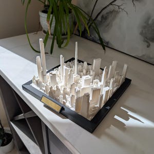 Peut inclure: Une maquette architecturale blanche imprimée en 3D représentant une ligne d'horizon urbaine avec de nombreux gratte-ciel de hauteurs variées, montée dans un cadre carré noir incluant une petite plaque rectangulaire dorée vierge sur le bord avant.