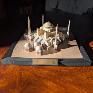 Può includere: Un modello architettonico in legno chiaro della Basilica di Santa Sofia con una cupola centrale, quattro minareti e strutture circostanti più piccole, montato su una base piatta con cornice nera e una targhetta rettangolare vuota in metallo dorato.