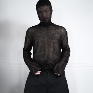 May include: A person wearing a black, sheer turtleneck shirt and matching black pants. The person is also wearing a black mesh hood that covers the head and face. The outfit is set against a plain white background.