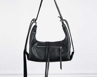 May include: A black shoulder bag with a curved top silhouette, featuring a horizontal zippered pocket on the front with two long, thin leather pull tabs. The bag includes a long, adjustable shoulder strap attached with silver-toned metal hardware.