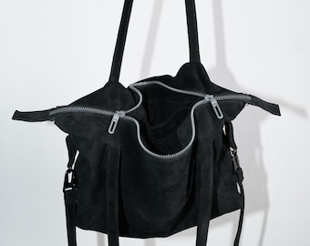 May include: Black suede tote bag with a silver zipper closure. The bag has long, thin straps for carrying over the shoulder or by hand. The bag is open, revealing the interior. The bag is hanging against a white background.