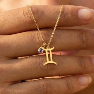 May include: A gold necklace with a Gemini zodiac symbol pendant and a small purple gemstone. The necklace is held up in front of a person's face. The pendant is a stylized representation of the Gemini constellation.