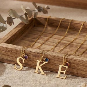 May include: Gold-colored initial necklaces with gemstone accents displayed on a wooden tray. The letters S, K, and E are visible, each with a small gemstone. A gold ring sits on a small white dish. The background includes decorative pottery and dried flowers.