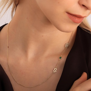 May include: A delicate gold necklace with a pendant featuring the letter "S", a green gemstone, and a decorative circular design. The necklace is worn on a person's neck, against a dark top.