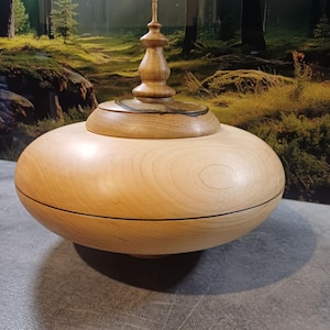 May include: A light-colored wooden bowl with a lid. The bowl has a rounded shape with a dark line around the middle. The lid is topped with a decorative finial. The wood grain is visible.