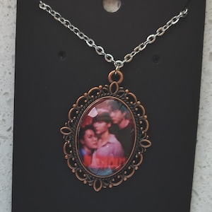 May include: A silver chain necklace with an oval pendant. The pendant has a bronze-colored filigree frame and a photo of people. The necklace is displayed on a black card.