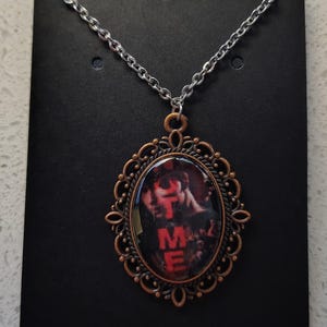 May include: A silver-toned chain necklace with a copper-colored filigree pendant. The oval pendant features a red and black image with the letters "TME" in red. The necklace is displayed on a black card.