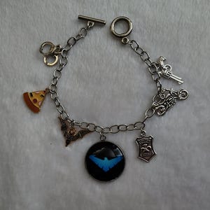 May include: Silver charm bracelet with various charms, including a slice of pizza, handcuffs, a bat, a shield, a gun, a motorcycle, and a black pendant with a blue emblem. The bracelet has a toggle clasp.