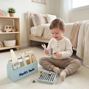 May include: A light blue wooden toolbox with the text "Noah's Tools" contains toy tools, including a saw, wrench, hammer, and ruler. A child is playing with the toy tools and a peg board with wooden pegs. The tools are light blue and natural wood.
