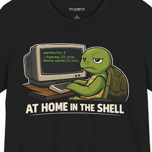 May include: Black t-shirt featuring a cartoon turtle at a computer. The turtle has a grumpy expression and is typing on a keyboard. The computer screen displays code. The text "AT HOME IN THE SHELL" is printed below the turtle.