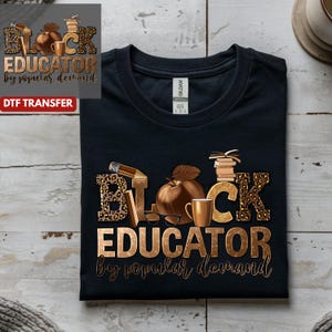 May include: Black t-shirt with a gold and leopard print design that reads "BLACK EDUCATOR by POPULAR DEMAND." The design includes a pencil, apple, coffee cup, books, and glasses. The shirt is made of a soft material.