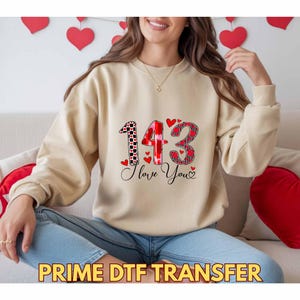 May include: A beige sweatshirt featuring the numbers "143" in a checkered and red heart design, with the words "I love You" in script. Small red hearts are scattered around the numbers. The text "PRIME DTF TRANSFER" is at the bottom.