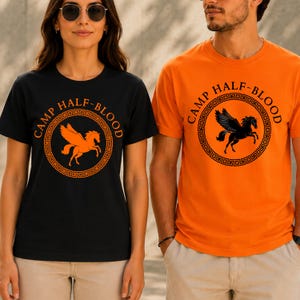May include: Black and orange t-shirts with the text "CAMP HALF-BLOOD" in a circular design. The black shirt features an orange pegasus, while the orange shirt has a black pegasus. Both shirts have a Greek key border.