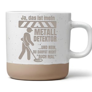 Könnte beinhalten: Ein weißer Keramikbecher mit Sprenkeloptik und beigem Boden. Auf der Vorderseite befindet sich eine Grafik einer Person mit einem Metalldetektor und der Text: Ja, das ist mein METALL DETEKTOR ...UND NEIN, DU DARFST NICHT "AUCH MAL". Mit seitlichem Henkel.