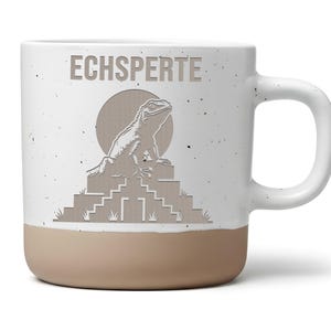 Könnte beinhalten: Ein weißer Keramikbecher mit gesprenkelter Oberfläche und beigem Boden. Die Vorderseite zeigt eine beige Grafik einer Eidechse auf einer Stufenpyramide vor einer kreisförmigen Sonne, mit dem darüber gedruckten Schriftzug ECHSPERTE in Beige.