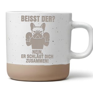 Könnte beinhalten: Ein weißer Keramikbecher mit gesprenkelter Oberfläche und beigem Boden. Auf der Vorderseite ist eine Grafik einer Französischen Bulldogge mit Boxhandschuhen abgebildet, darüber und darunter der Text "BEISST DER? NEIN, ER SCHLÄGT DICH ZUSAMMEN!" in Beige.