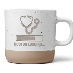 Könnte beinhalten: Eine gesprenkelte, weiße Keramik-Tasse mit beigefarbenem Boden und weißem Henkel. Die Tasse zeigt ein Stethoskop und eine Ladebalken-Grafik mit dem Text "DOCTOR LOADING..." in Grau.
